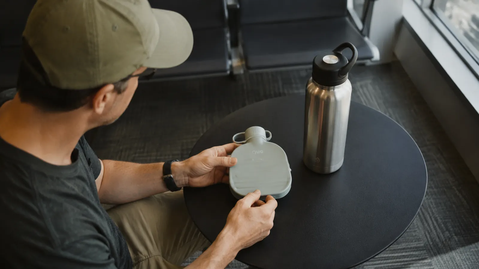 A collapsed water bottle and a hard-sided steel bottle side by side on an airport gate bench, next to a small carry-on backpack